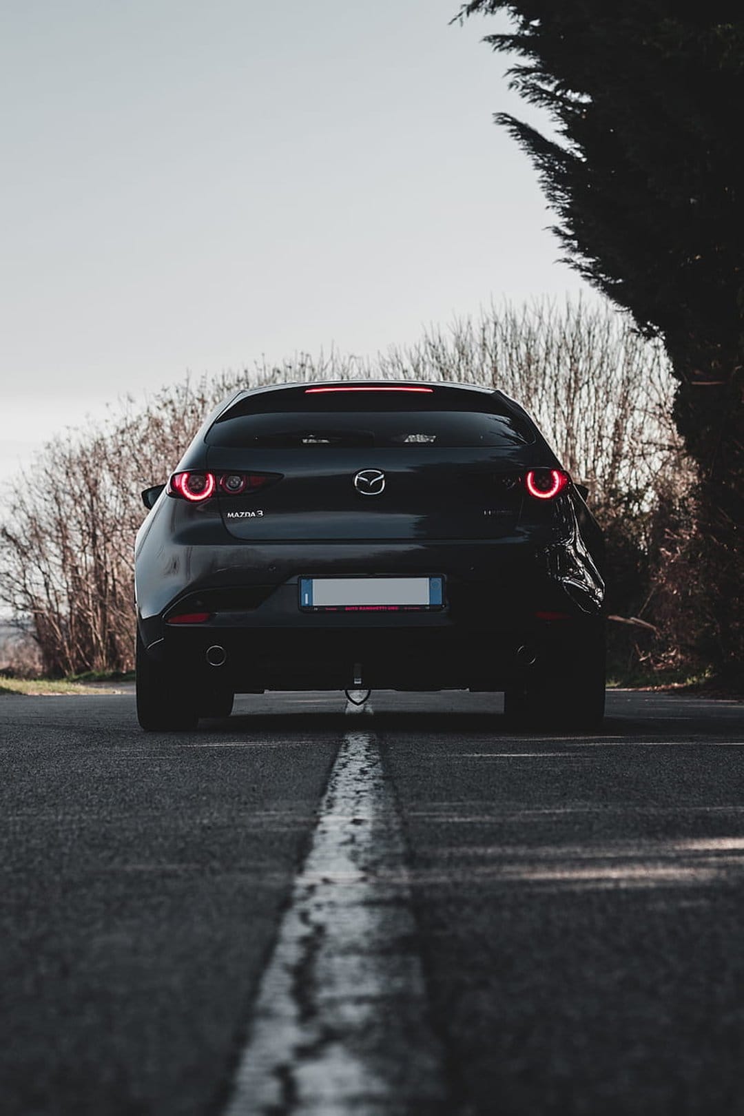 Black Mazda 3 hatchback rear view on an asphalt road with glowing red taillights and leafless autumn trees.