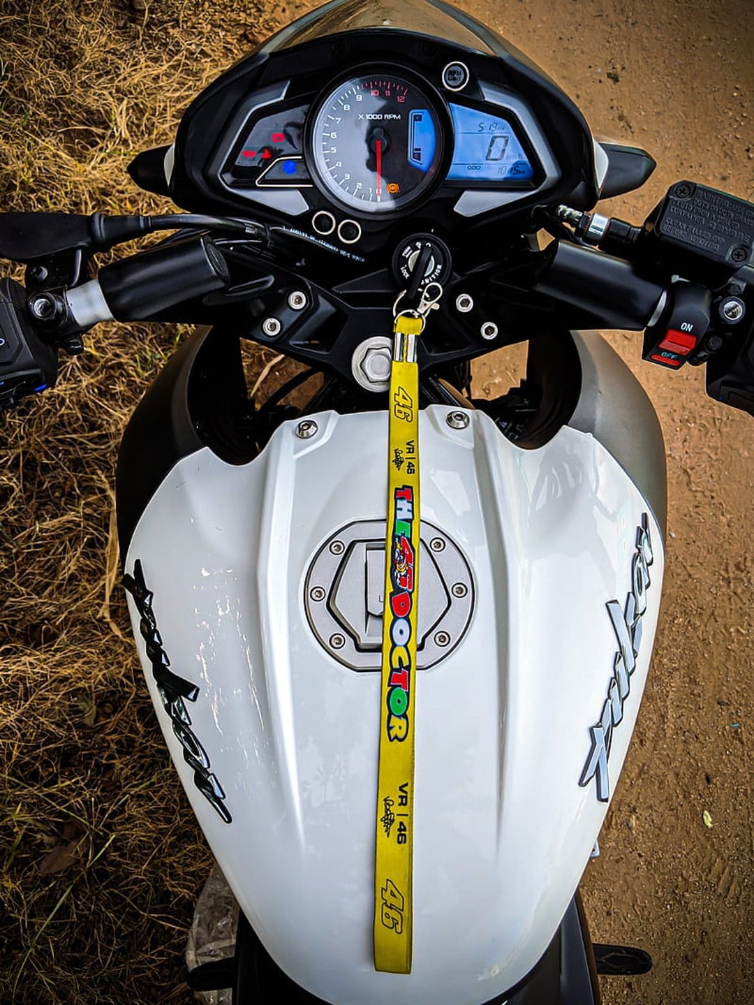 Motorcycle dashboard with a yellow VR46 lanyard hanging near a white fuel tank and digital tachometer on grass.