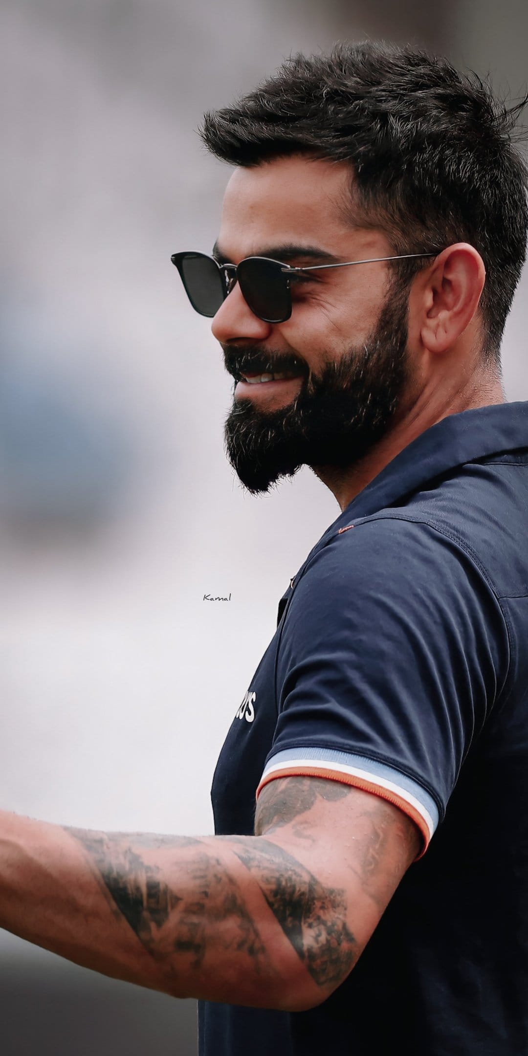 Virat Kohli smiles in profile wearing dark sunglasses and a navy blue polo shirt with visible arm tattoos.