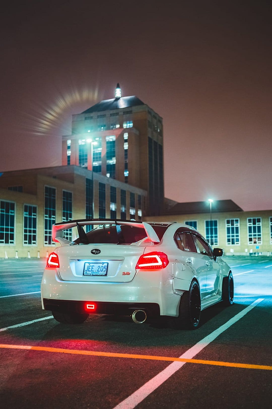 White Subaru WRX with widebody fenders and a large wing parked in a night lot with red taillights glowing.