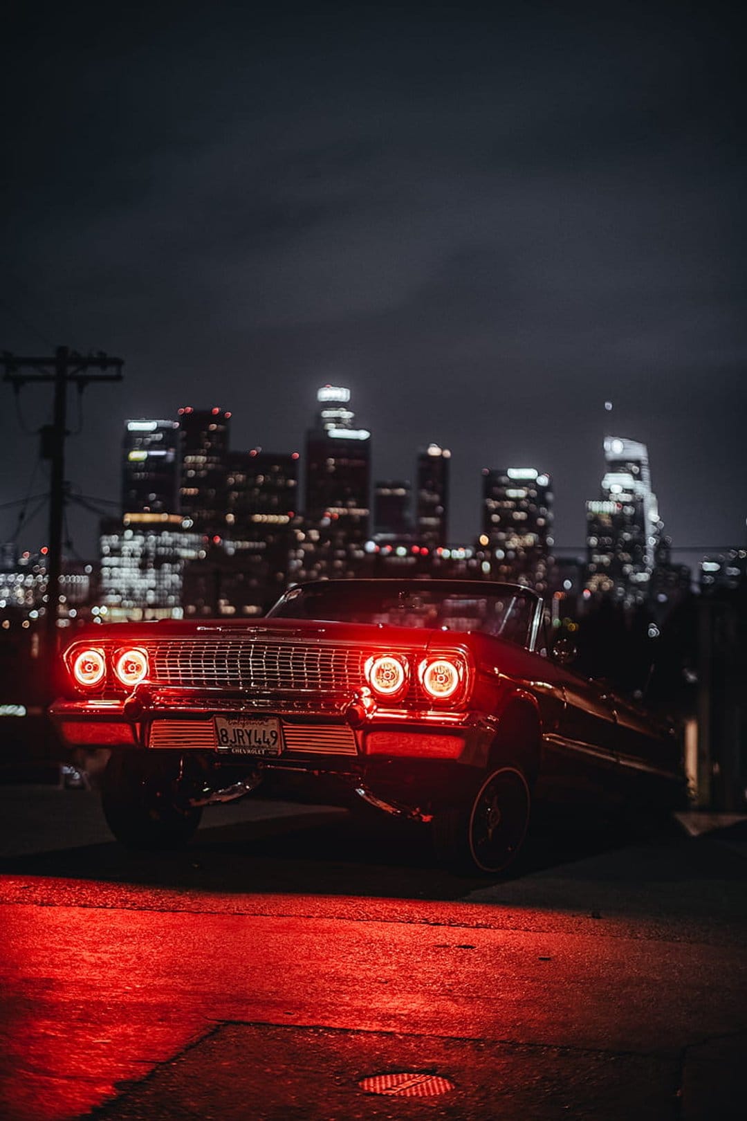 Red lowrider convertible parked under crimson streetlights with a glowing city skyline in the background.