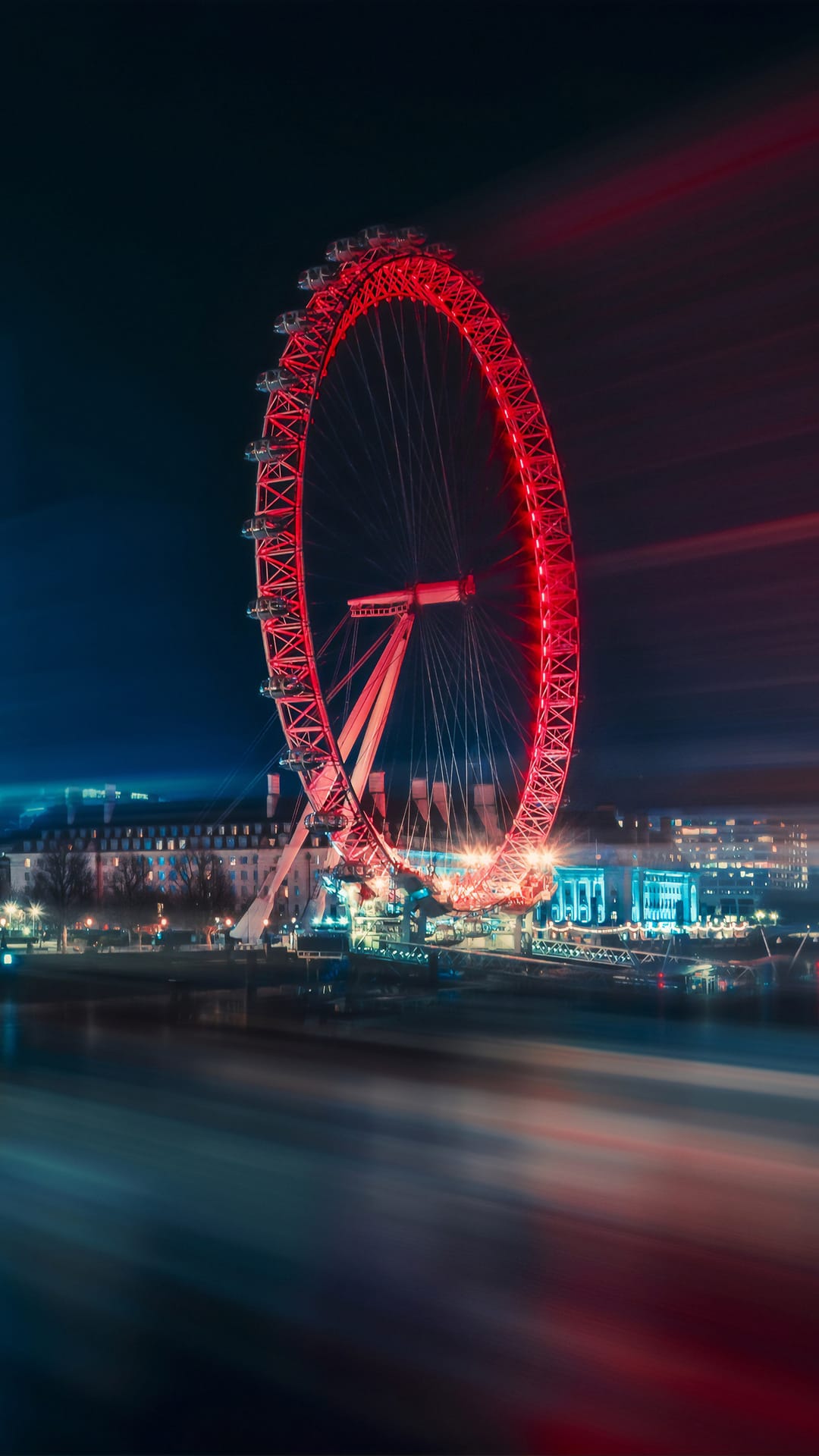 London Eye ferris wheel illuminated in red at night over the River Thames with long exposure light trails.