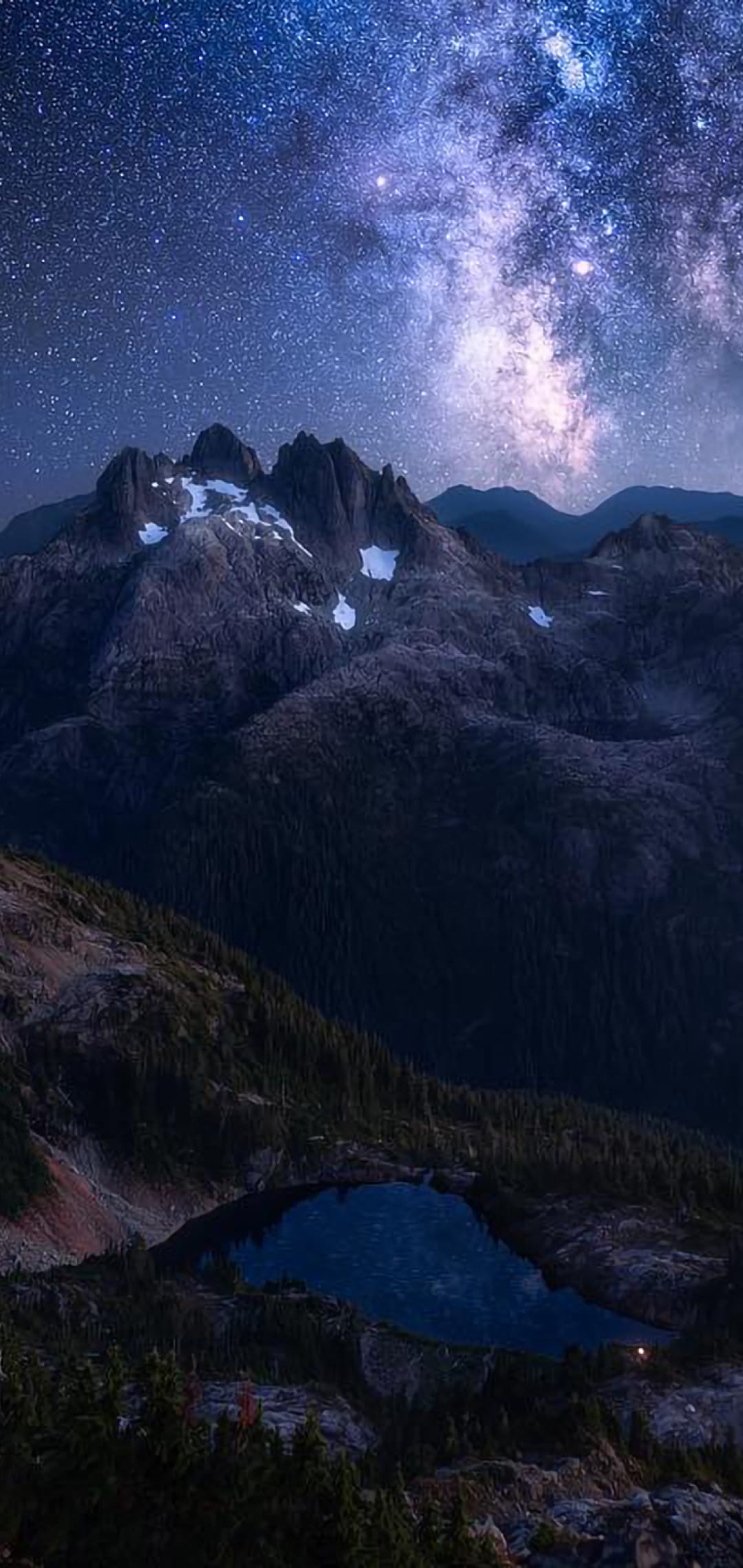 Milky Way galaxy core arcs above snow-dusted mountain peaks and a calm lake reflection in a vertical night scene.