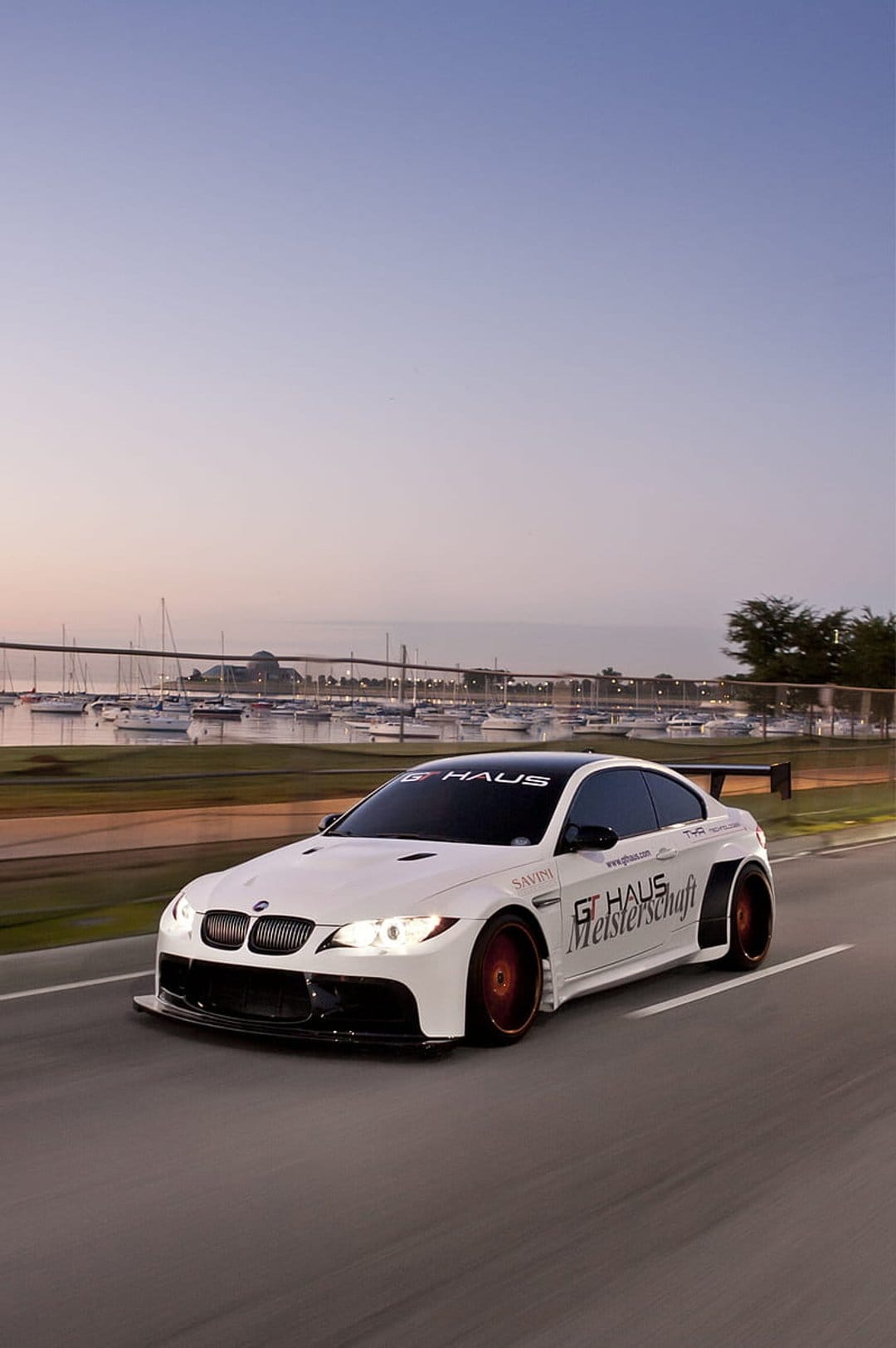 White BMW M3 with widebody kit and red wheels driving past sailboats during a vibrant coastal sunset.
