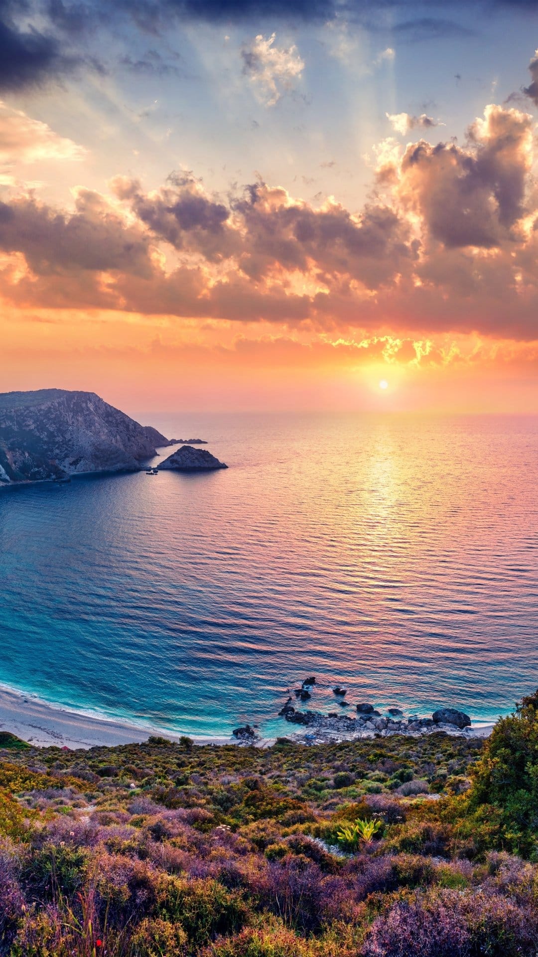Golden sun glows over a tropical coastline with purple plants, white sand, and turquoise ocean water seen from above.