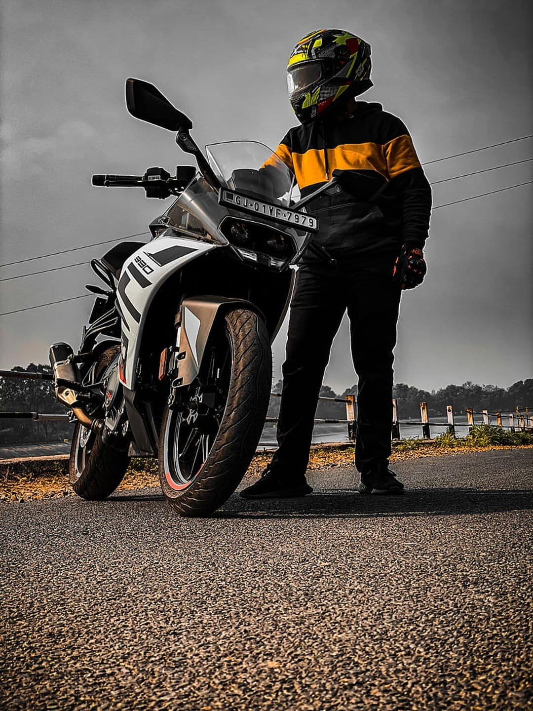 Biker in a colorful helmet stands by a white and black sportbike on a gray asphalt road under a cloudy sky.