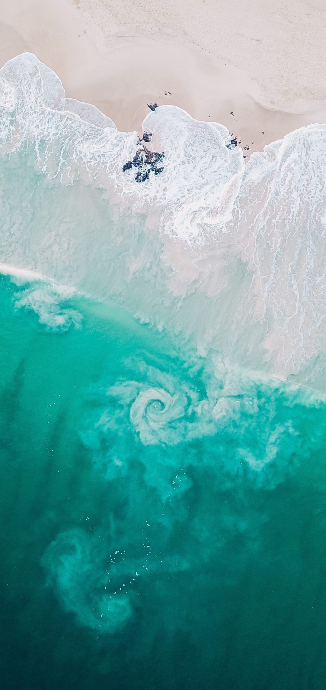Turquoise ocean waves with white foam patterns swirl against a sandy tropical shoreline from an aerial view.