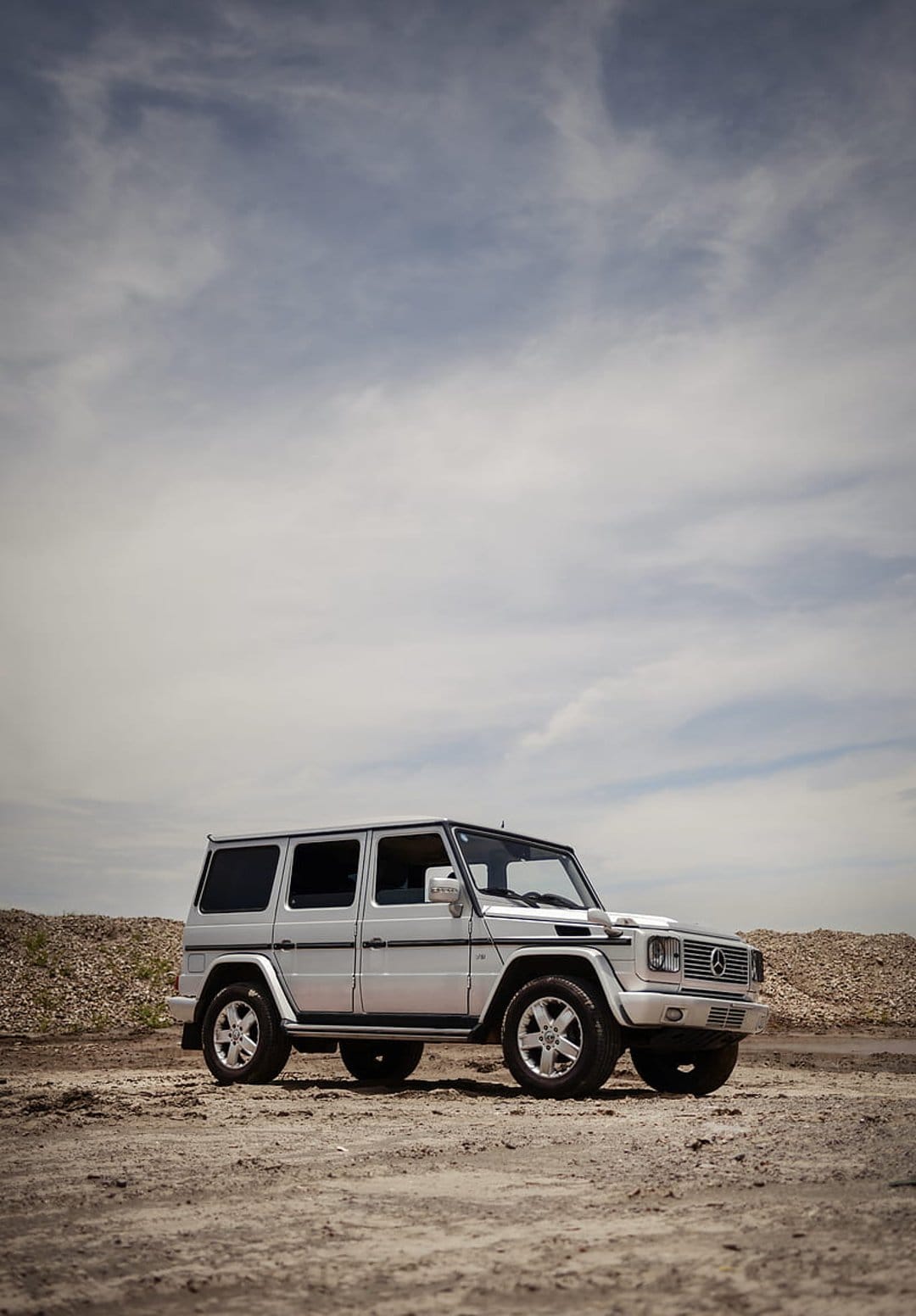 Silver Mercedes G-Class SUV parked on a gray gravel lot under an overcast sky with a boxy off-road profile.