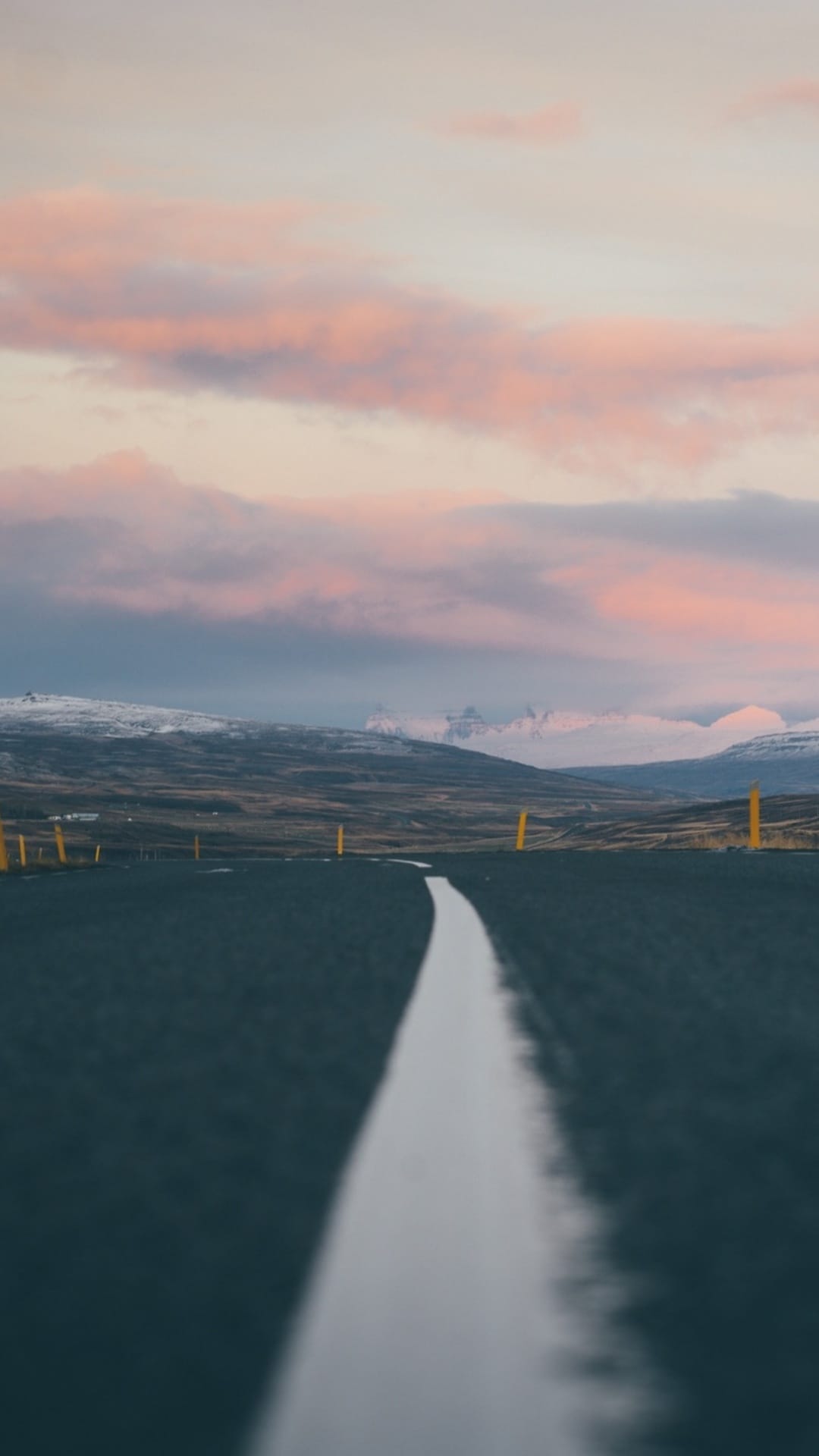Straight asphalt road with a white center line leading toward distant snow-covered mountains under a pink sky.