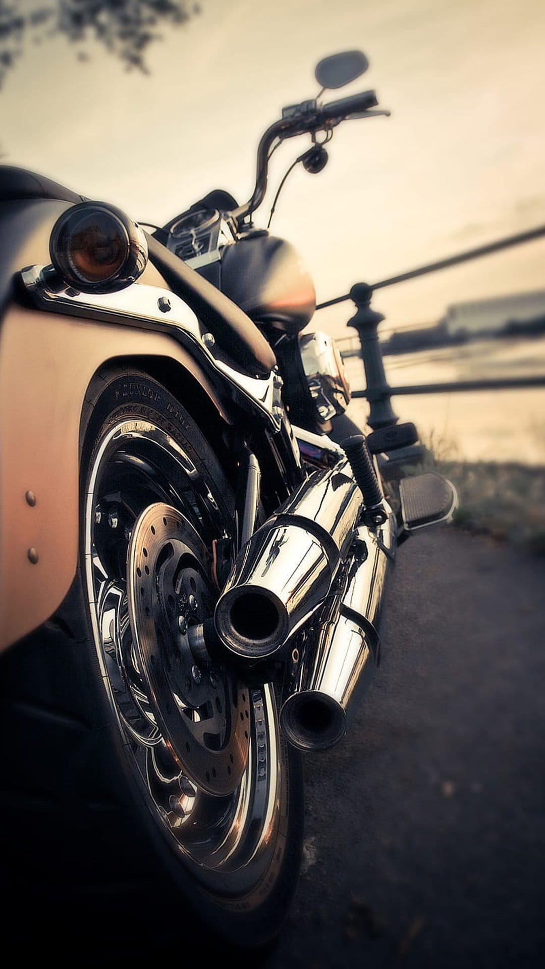 Chrome motorcycle exhaust pipes and a rear brake disc shine in a low-angle shot with a soft bokeh background.