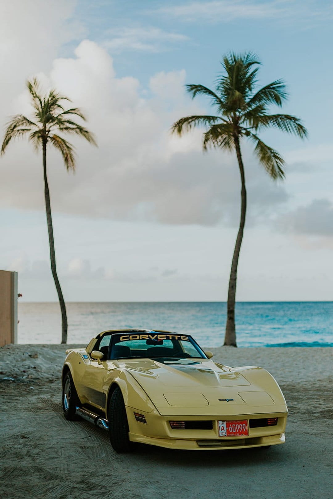 Yellow Chevrolet Corvette parked on a sandy beach between two palm trees with an ocean and cloudy sky background.