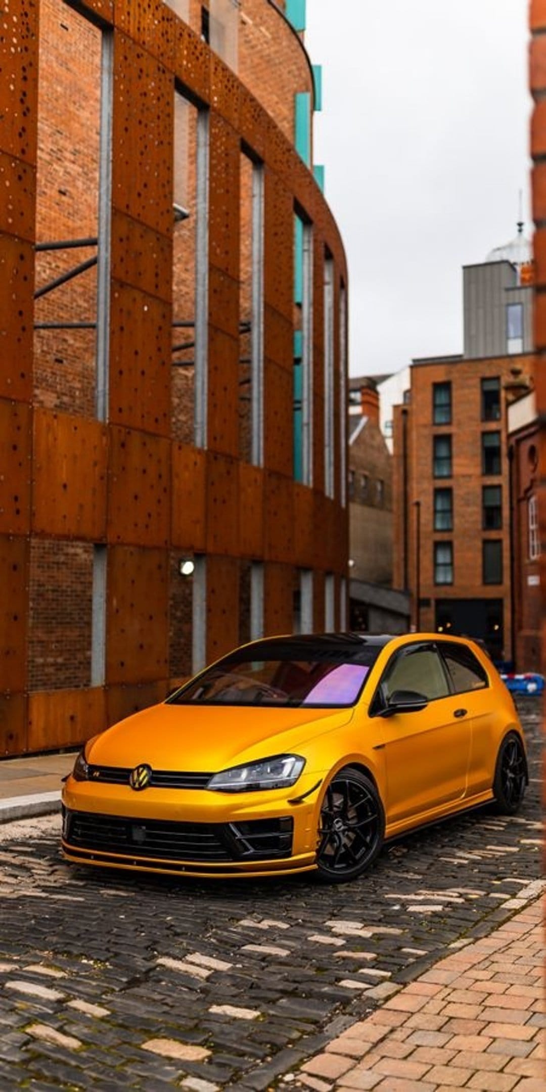Yellow Golf R hatchback on a cobblestone street with weathered metal and red brick industrial buildings behind.