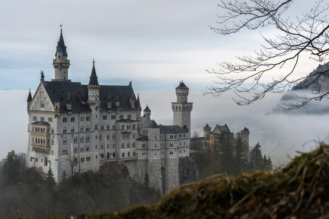 Neuschwanstein Castle with white stone towers perches on a rocky cliff surrounded by dense fog and winter trees.
