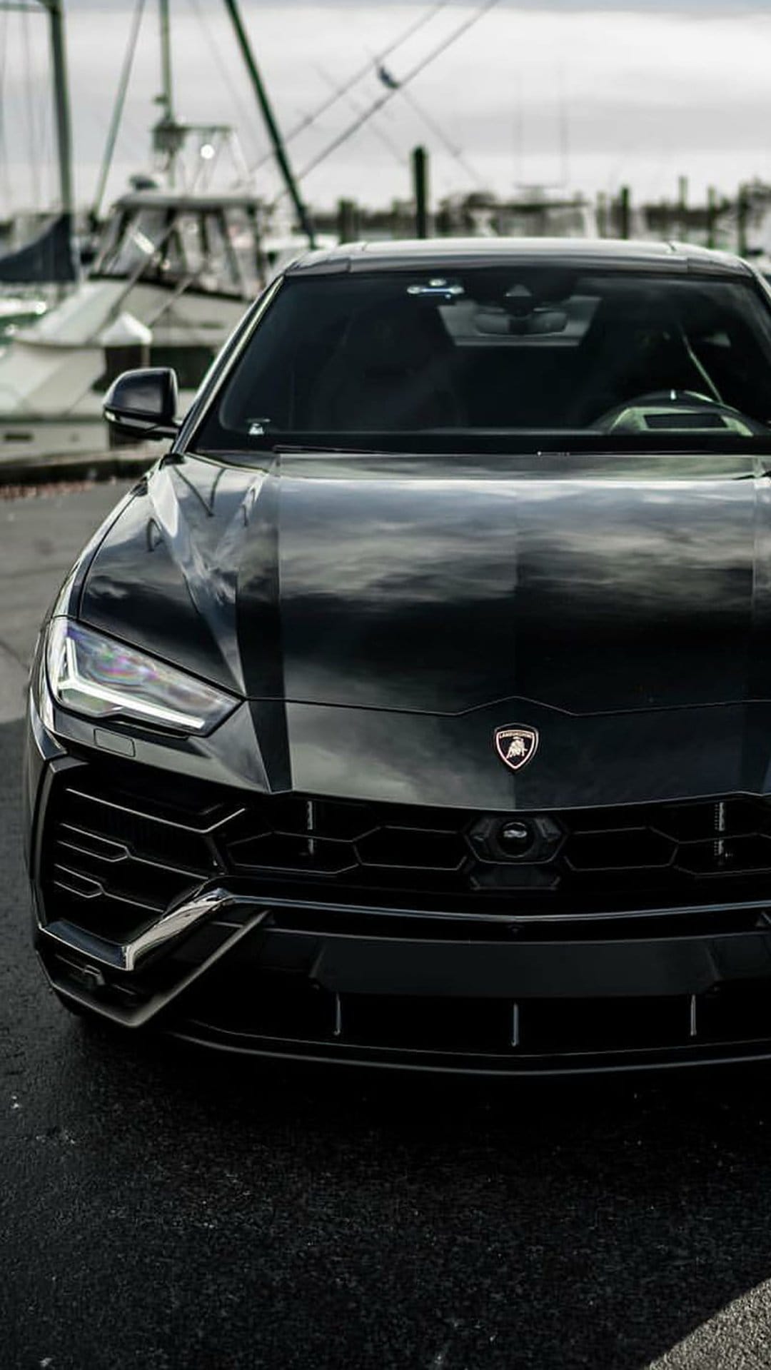 Black Lamborghini Urus SUV parked at a harbor with sailboats and a cloudy sky in the background for mobile.