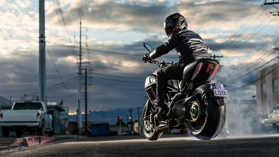 Black motorcycle performing a rear-tire burnout with thick white smoke on an urban street during a dramatic sunset.