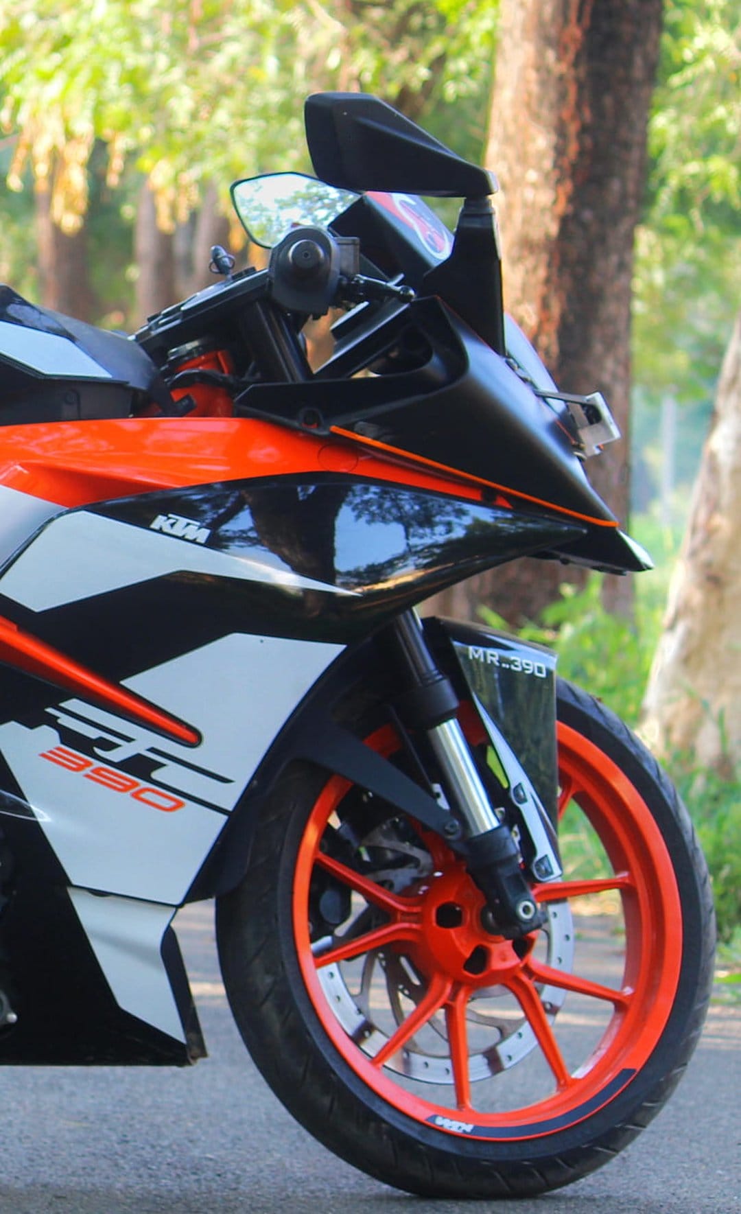 KTM RC 390 motorcycle orange front wheel and white fairing with a blurred green forest background in daylight.