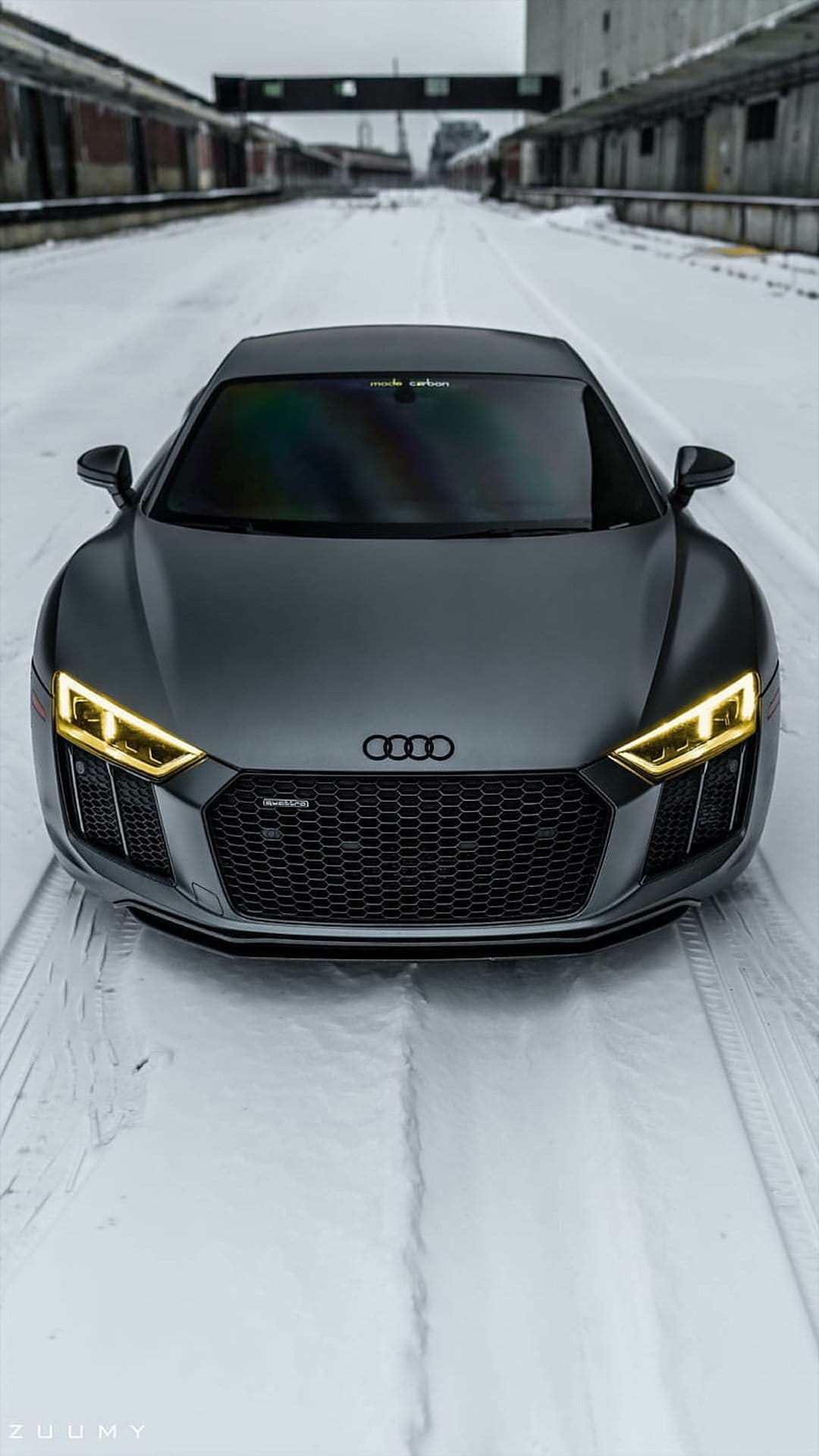Matte grey Audi R8 supercar parked on a snowy road with glowing yellow LED headlights and a blurred bridge.