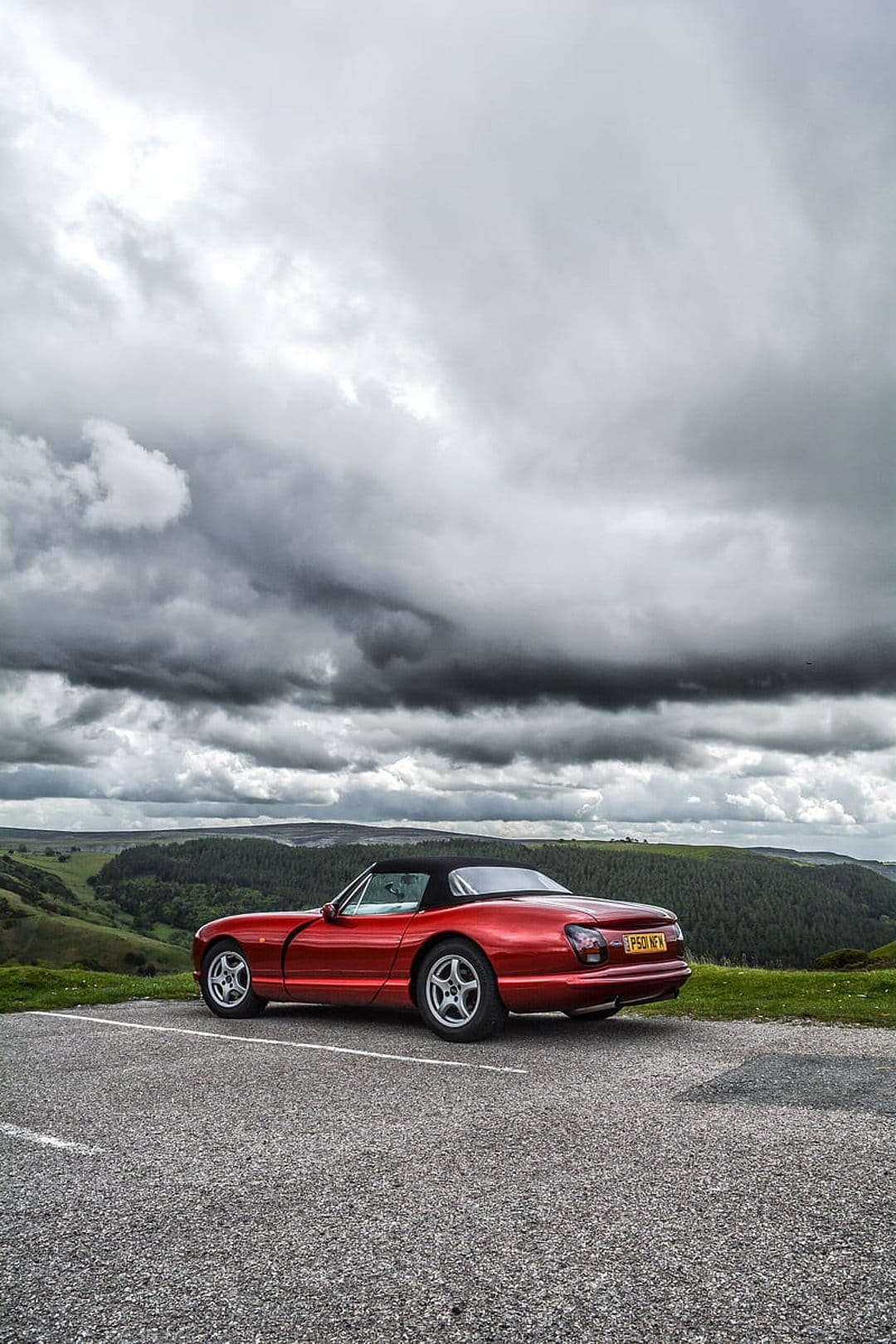 Red convertible sports car parked on gravel overlooking a green valley under dark, dramatic storm clouds.