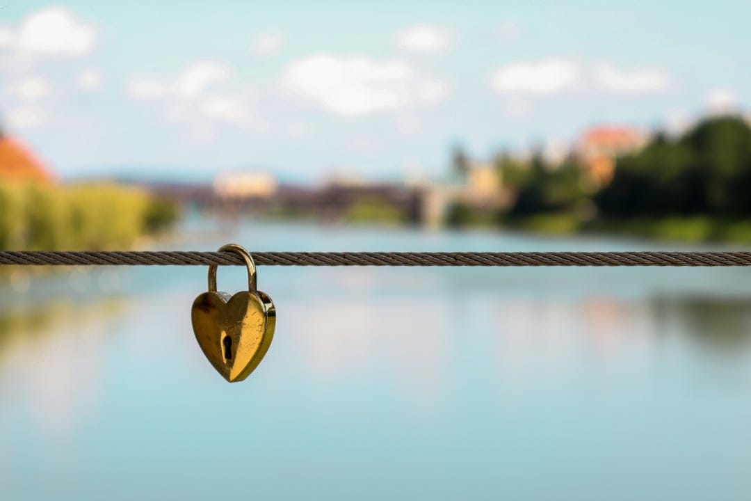 Golden heart-shaped lock secured to a steel bridge cable with a blurred river and cityscape in the background.