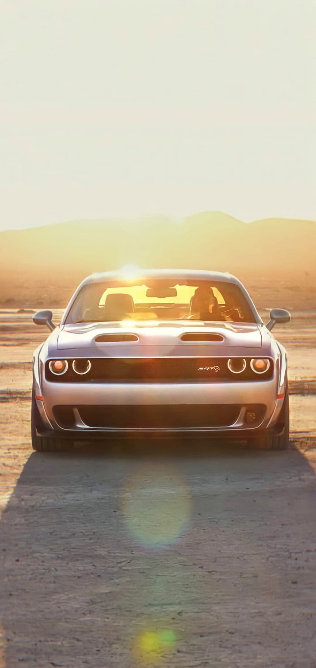 Silver Dodge Challenger Hellcat with glowing halo headlights parked on a desert road during a golden sunset.