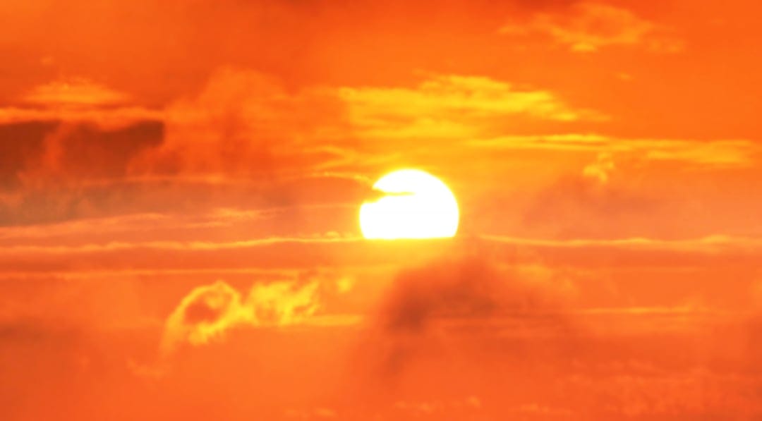 Bright white sun glows behind thin orange wispy clouds during a vibrant golden hour sunset in a clear sky.