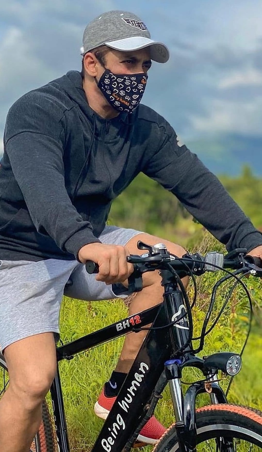 Man riding a black bicycle with orange accents in a grassy field while wearing a grey cap and a face mask.