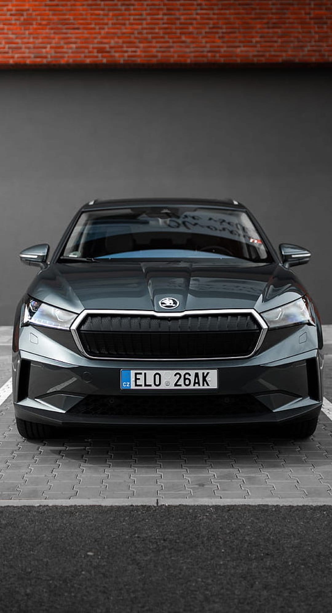 Skoda Enyaq iV electric SUV parked on pavement with a brick wall background and sharp front LED headlights.
