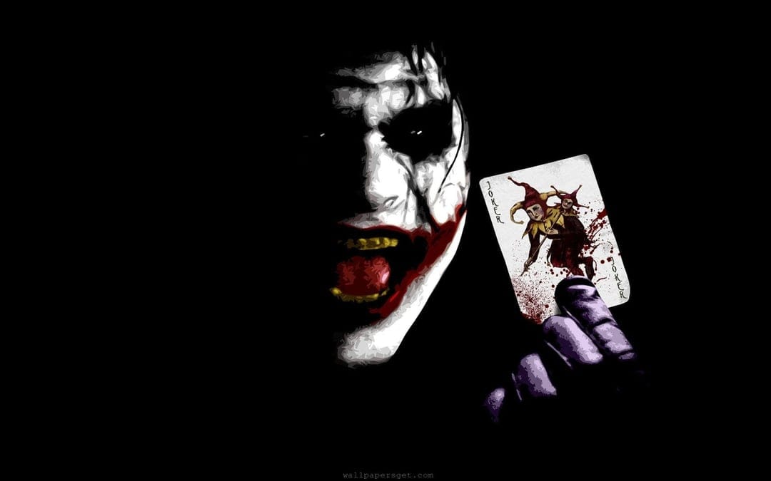 Joker with white face paint and a wide grin holds a blood-stained playing card against a dark black background.