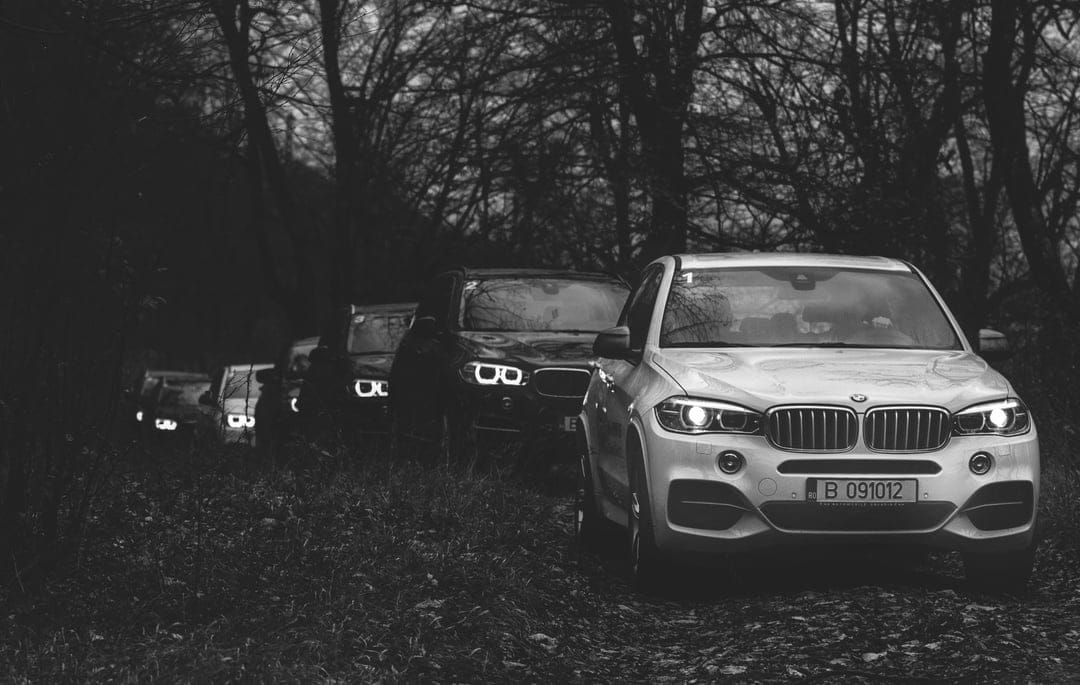Silver BMW X5 leads a line of SUVs through a dark, wooded forest trail in high-contrast black and white.