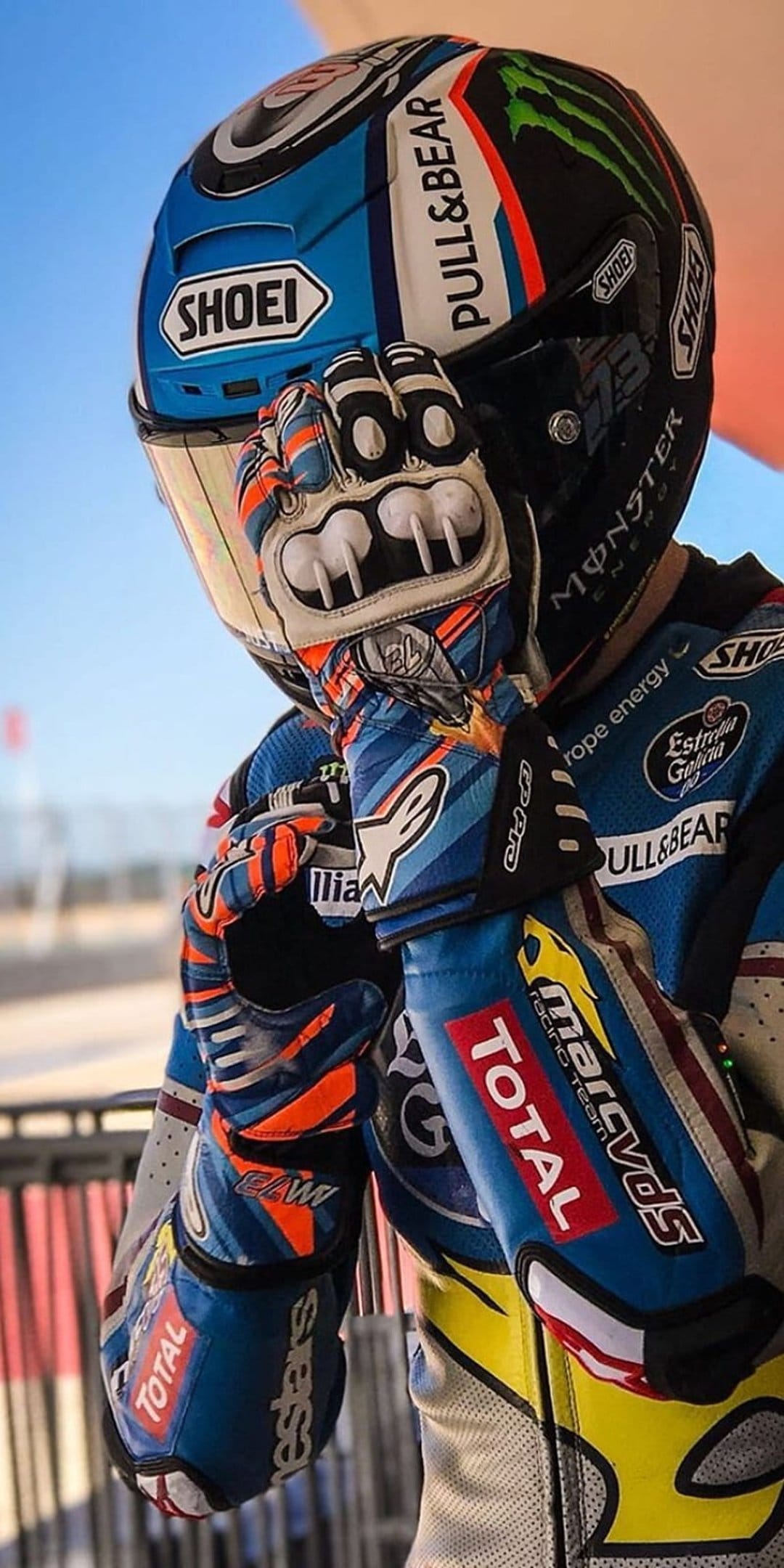 MotoGP racer in a blue Shoei helmet and orange Alpinestars gloves against a blurred outdoor track background.