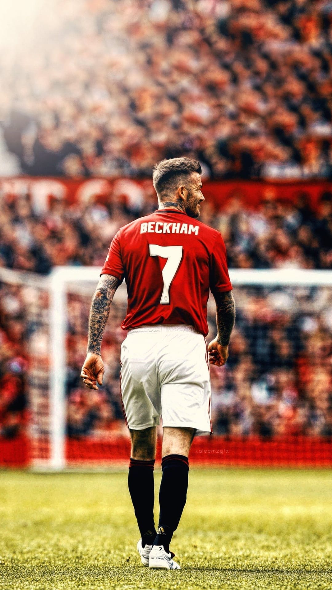 David Beckham walks across a grass pitch wearing a red Manchester United jersey with a white number 7 on his back.