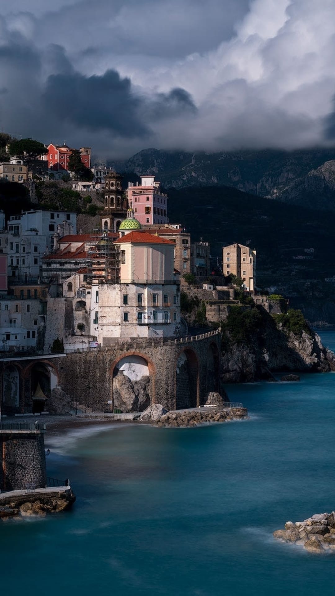 Pastel Italian houses on a rugged cliff overlook a turquoise sea under a dramatic dark sky with storm clouds.
