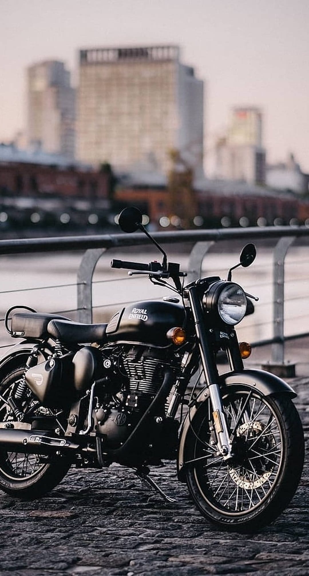 Black Royal Enfield motorcycle parked on cobblestones with a blurred urban waterfront and buildings behind it.