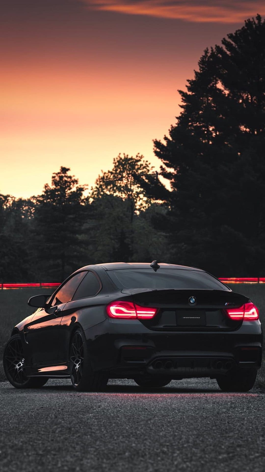Black BMW M4 parked on a gravel road with glowing red taillights under an orange and pink sunset sky.