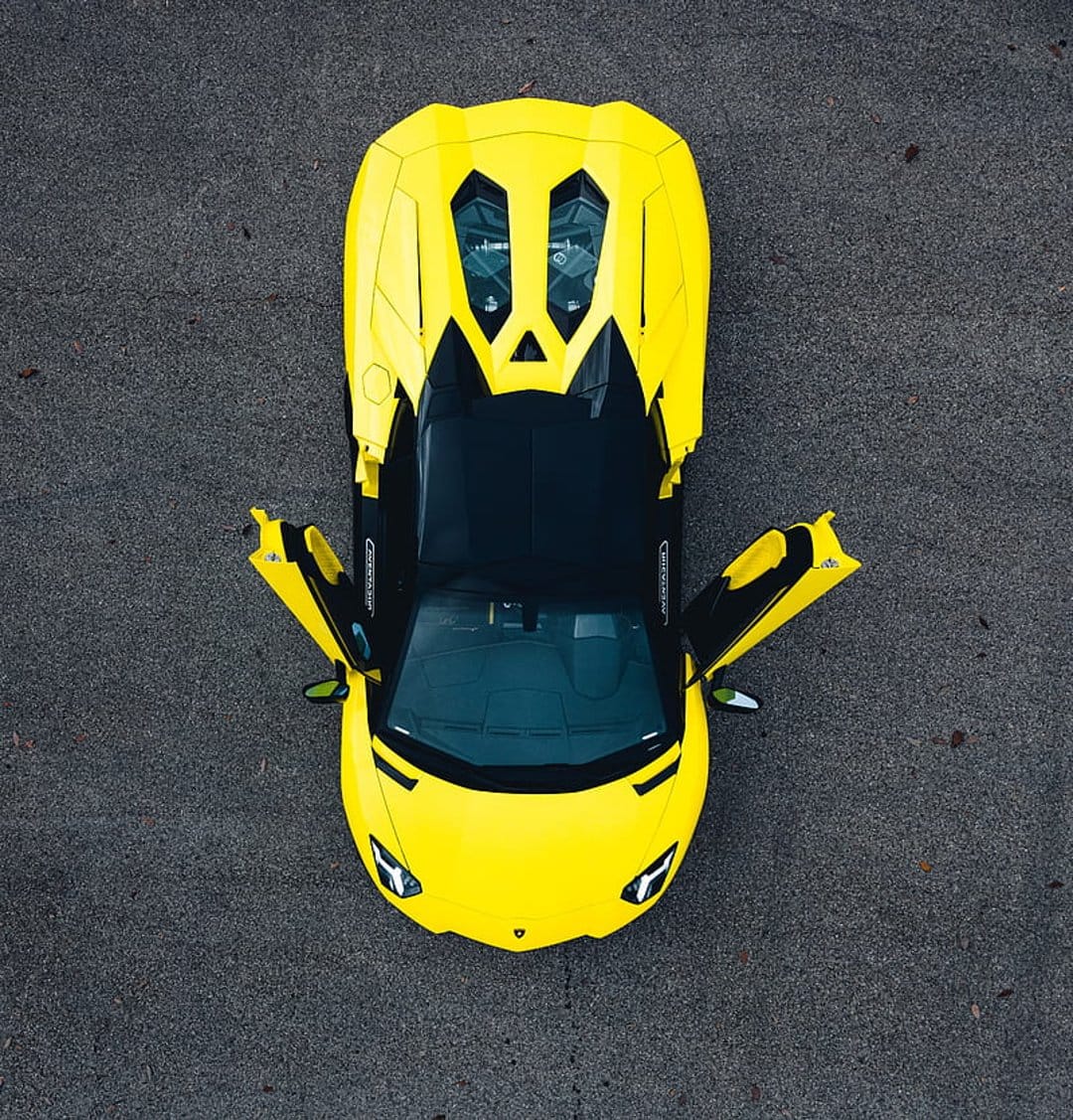 Yellow Lamborghini Aventador with open scissor doors parked on dark asphalt from a top-down aerial view.