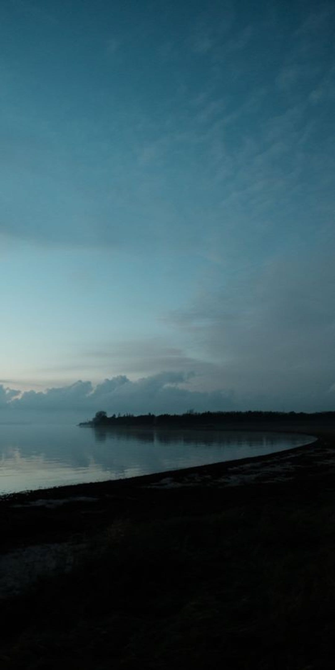 Misty coastline at dawn with calm water reflecting a soft blue sky and a dark silhouetted shore in the foreground.