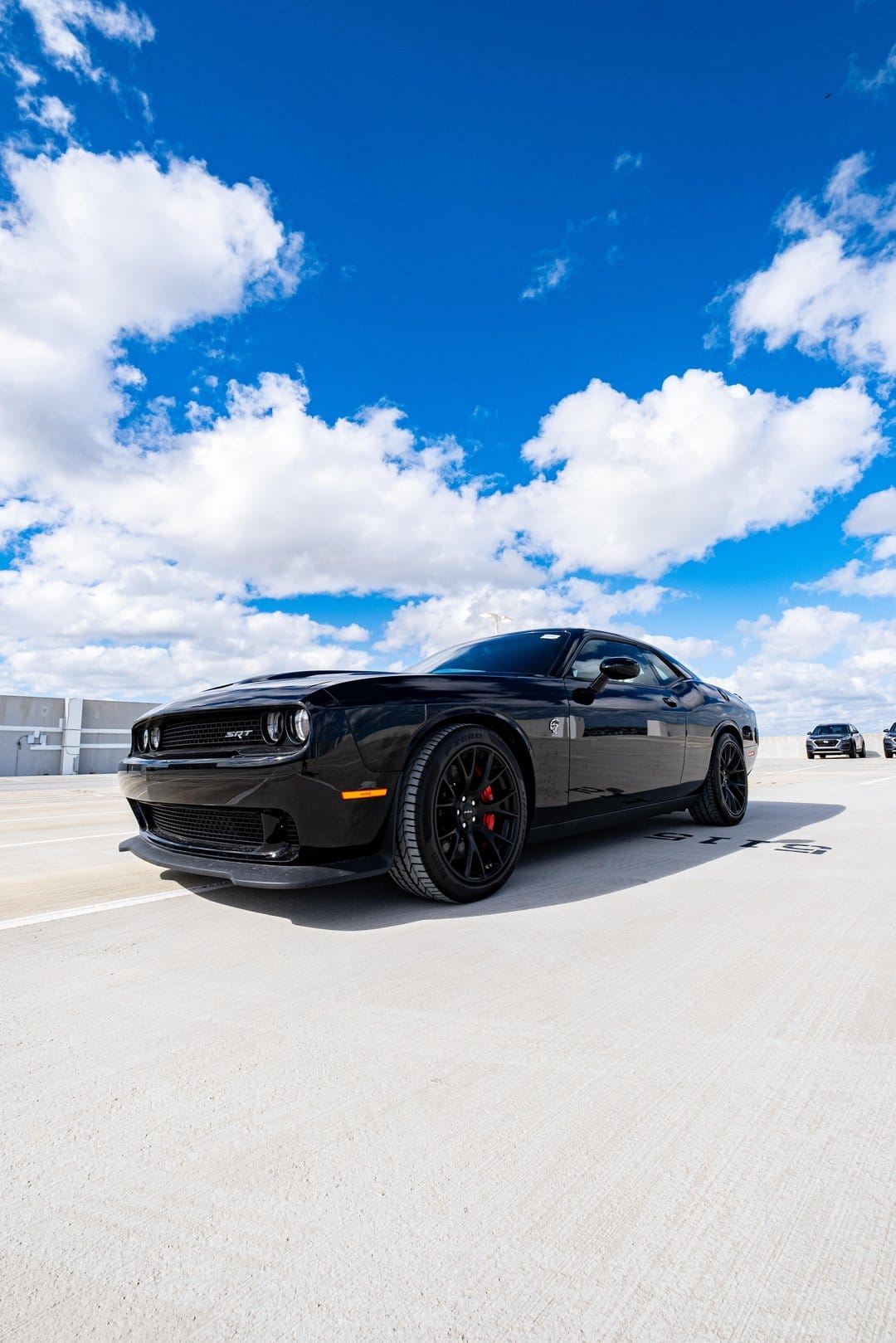 Black Dodge Challenger SRT Hellcat on a rooftop with red calipers, alloy wheels, and a bright blue cloudy sky.