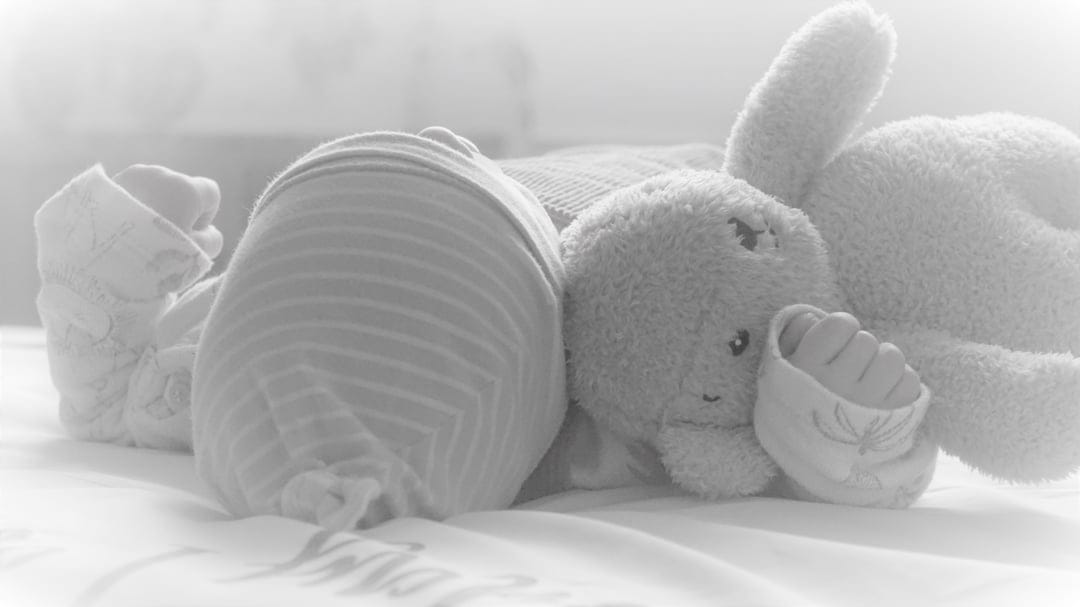 Sleeping infant in a striped onesie lying next to a soft plush bunny toy on a textured fabric surface.