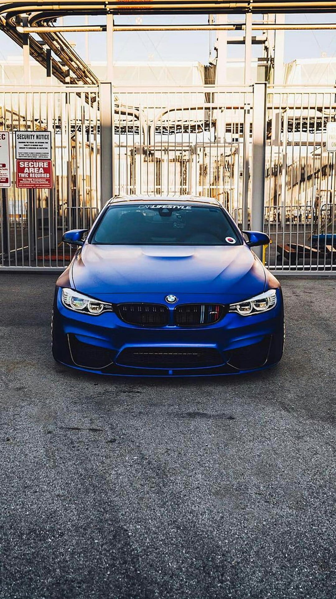Blue BMW M4 sports car parked in front of a gray metal security gate at an industrial facility front view.