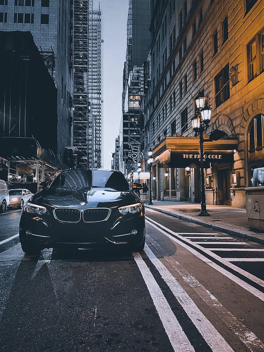 Black luxury car parked on wet asphalt at night, surrounded by illuminated skyscrapers and city buildings.