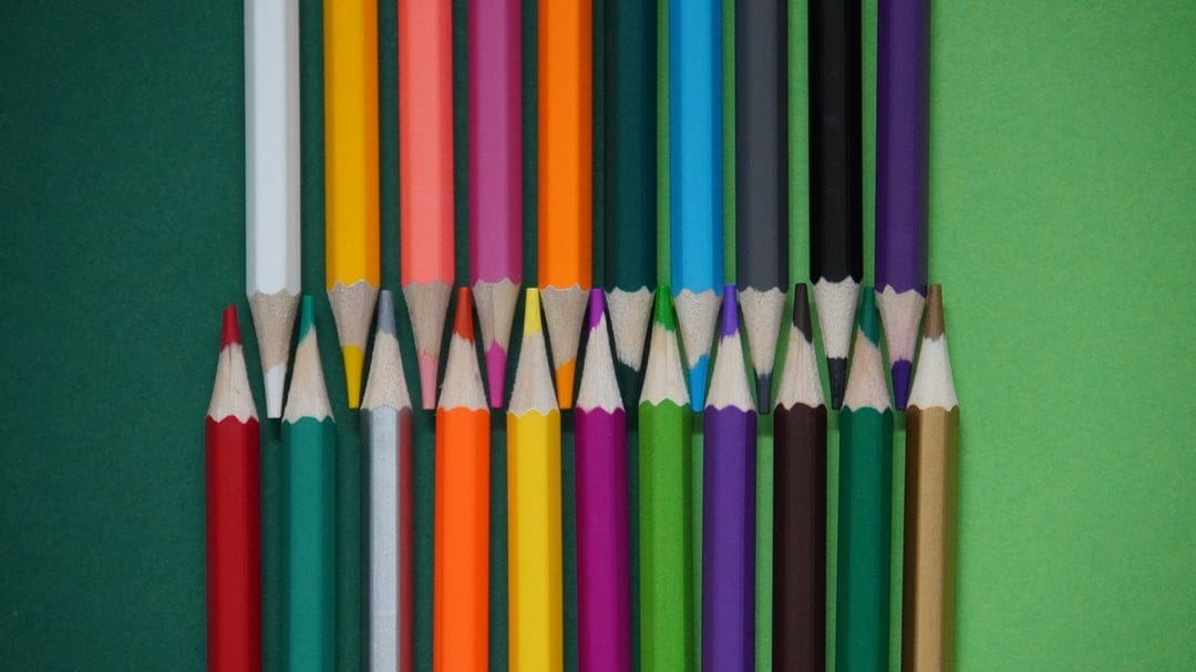 Rainbow-colored pencils form a precise V-shape on a split green background in a minimalist overhead shot.