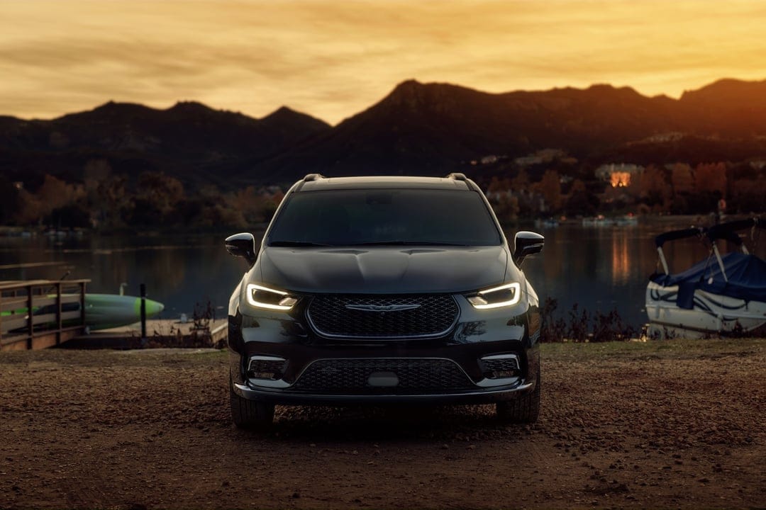 Black Chrysler Pacifica with glowing headlights parked by a calm lake against a mountain sunset background.