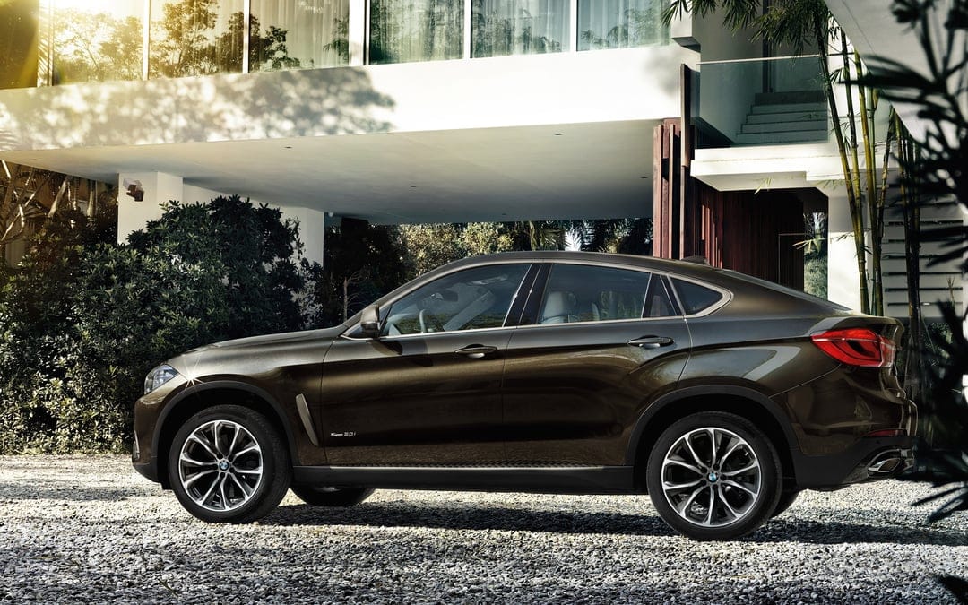 Bronze BMW X6 parked on a gravel driveway in front of a modern white house with golden sunlight flares.