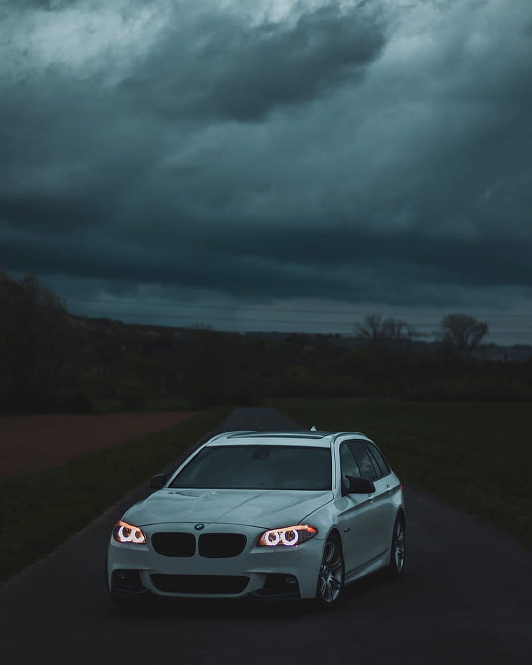 White BMW station wagon with glowing headlights parked on a dark highway under heavy gray storm clouds.