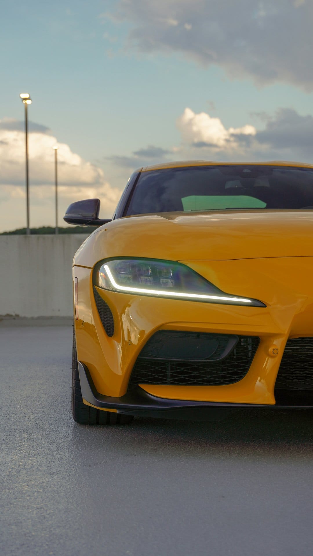 Yellow Toyota Supra headlight with glowing LEDs on asphalt under a cloudy sunset sky at a low camera angle.