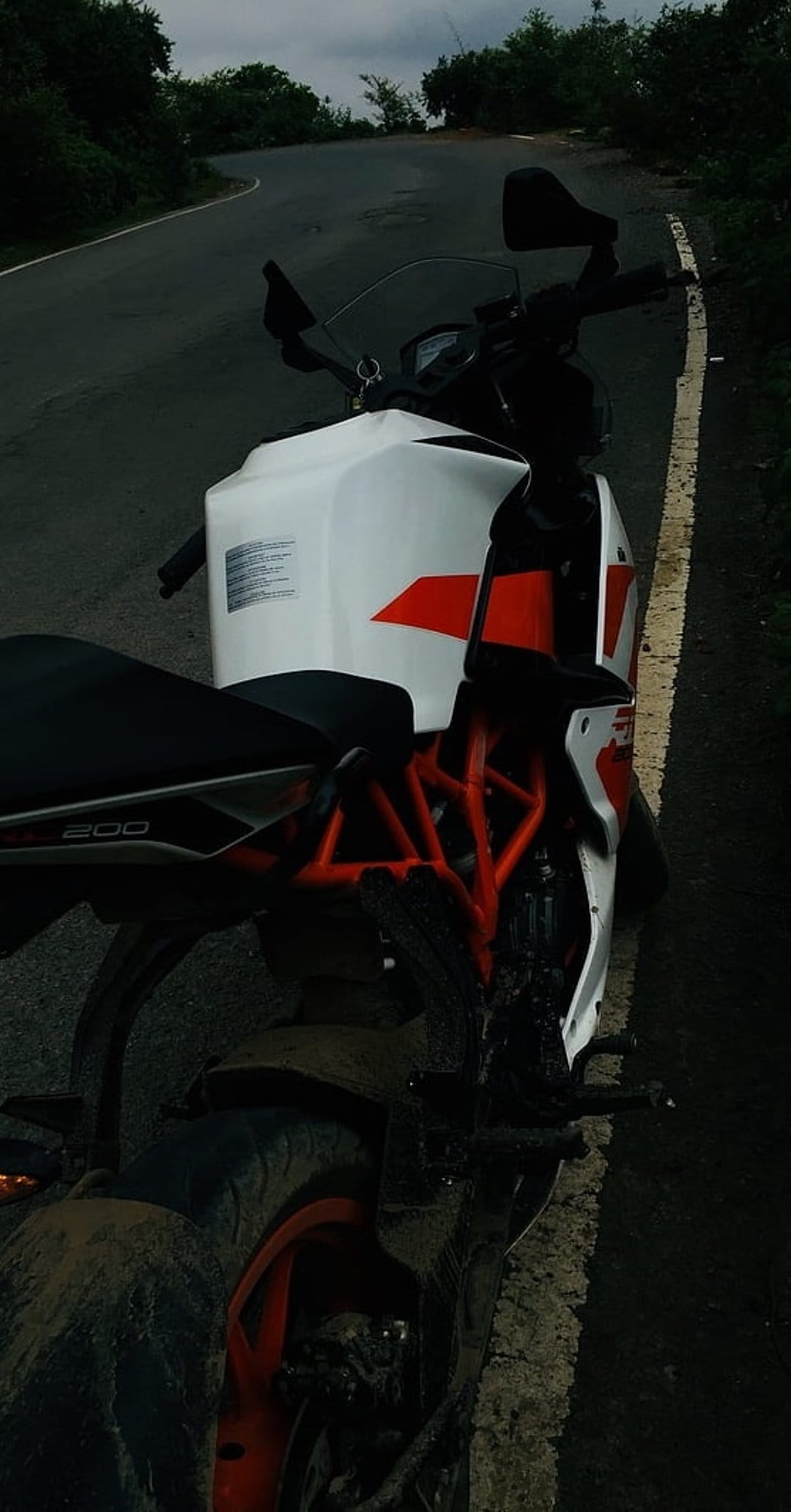 KTM RC 200 motorcycle with an orange frame parked on a wet, winding asphalt road under a cloudy, overcast sky.