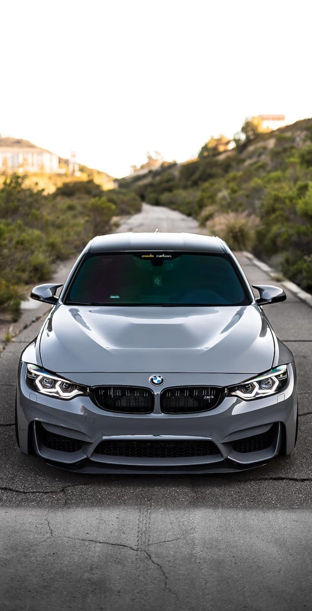BMW M4 in metallic grey parked on a cracked asphalt road with green rolling hills under a bright clear sky.