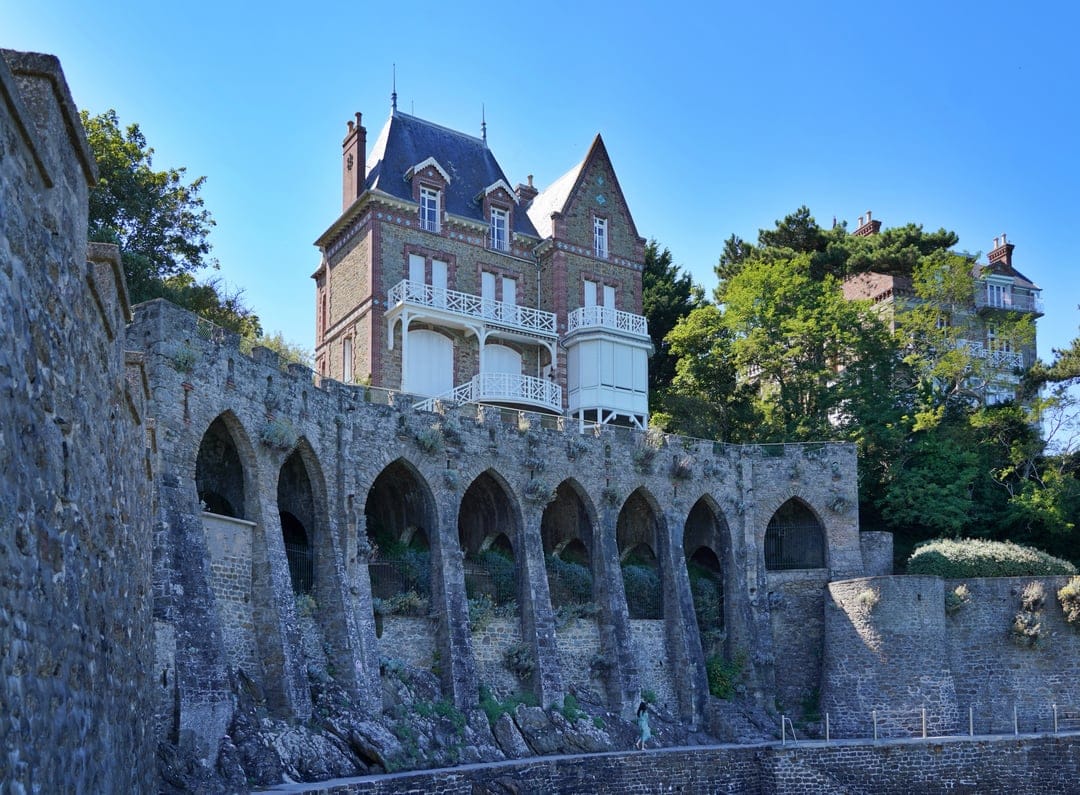 Gothic stone arches support a grand brick villa with white balconies under a clear blue sky with a lone pedestrian.