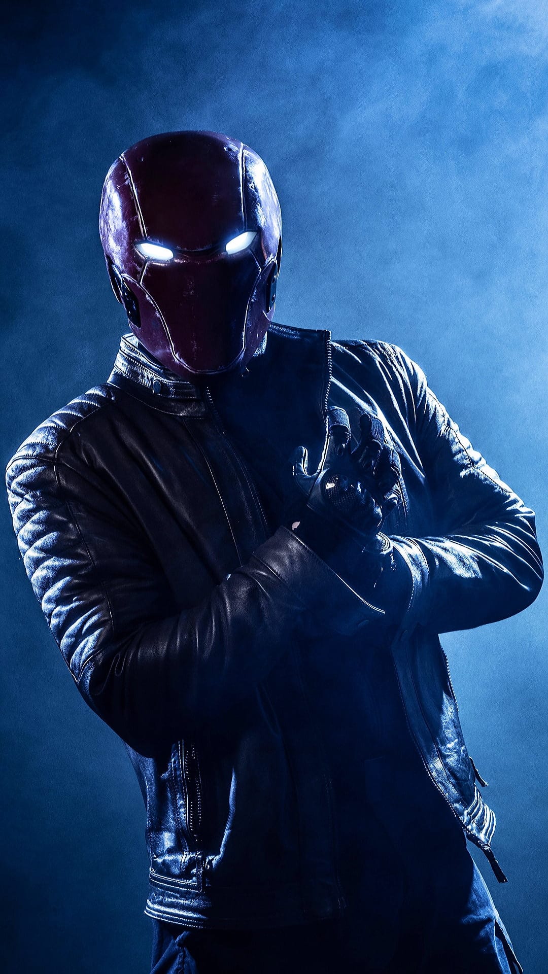 Red Hood wearing a crimson helmet and leather jacket stands before blue smoke with glowing white eyes.
