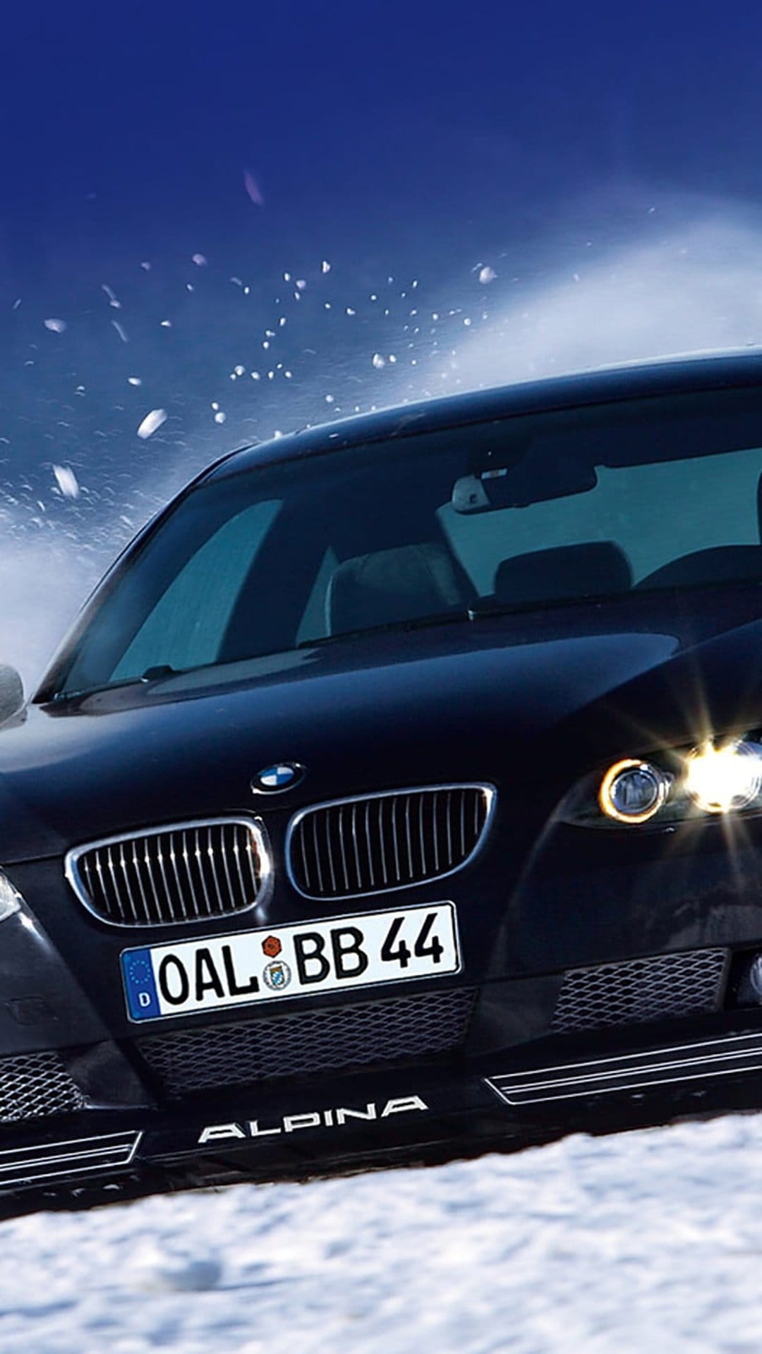 Alpina BMW sedan driving through thick snow with glowing headlights and frozen particles flying in the air.